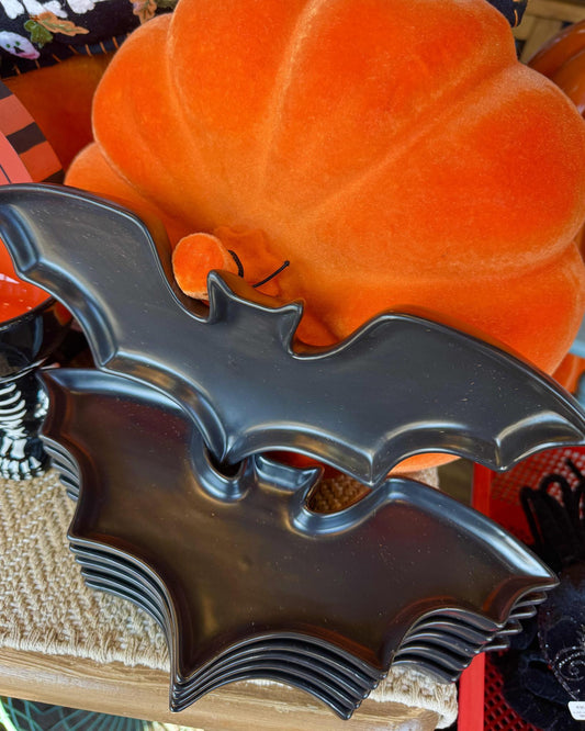A glossy black bat-shaped serving platter or tray displayed in front of a large orange pumpkin decoration.