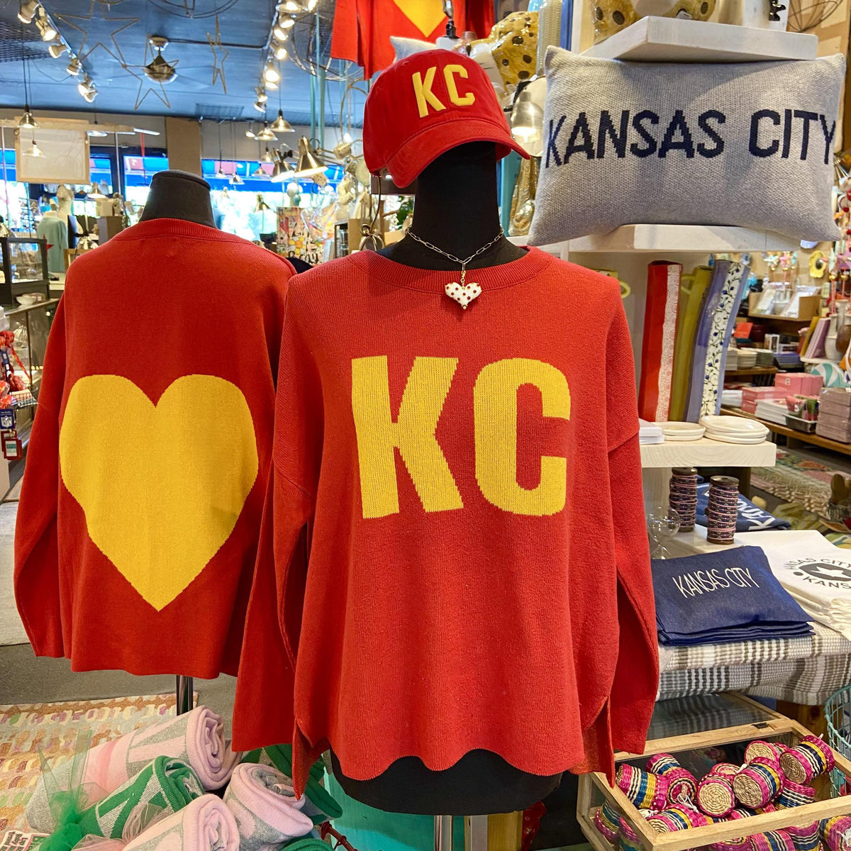 Red sweatshirts with 'KC' and heart designs on mannequins in a store setting.