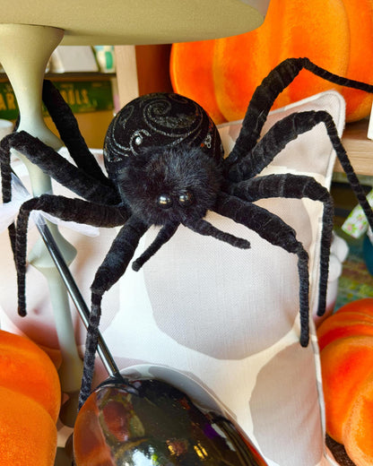 A black plush spider decoration with glittery details on its abdomen, posed on a white pillow with orange pumpkins in the background.