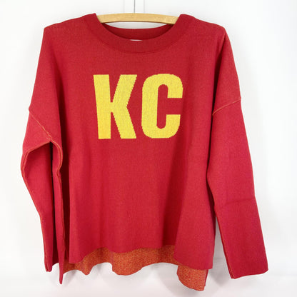 A red and gold pullover sweater with 'KC' text on the front and a heart shape on the back, displayed on a wooden hanger.