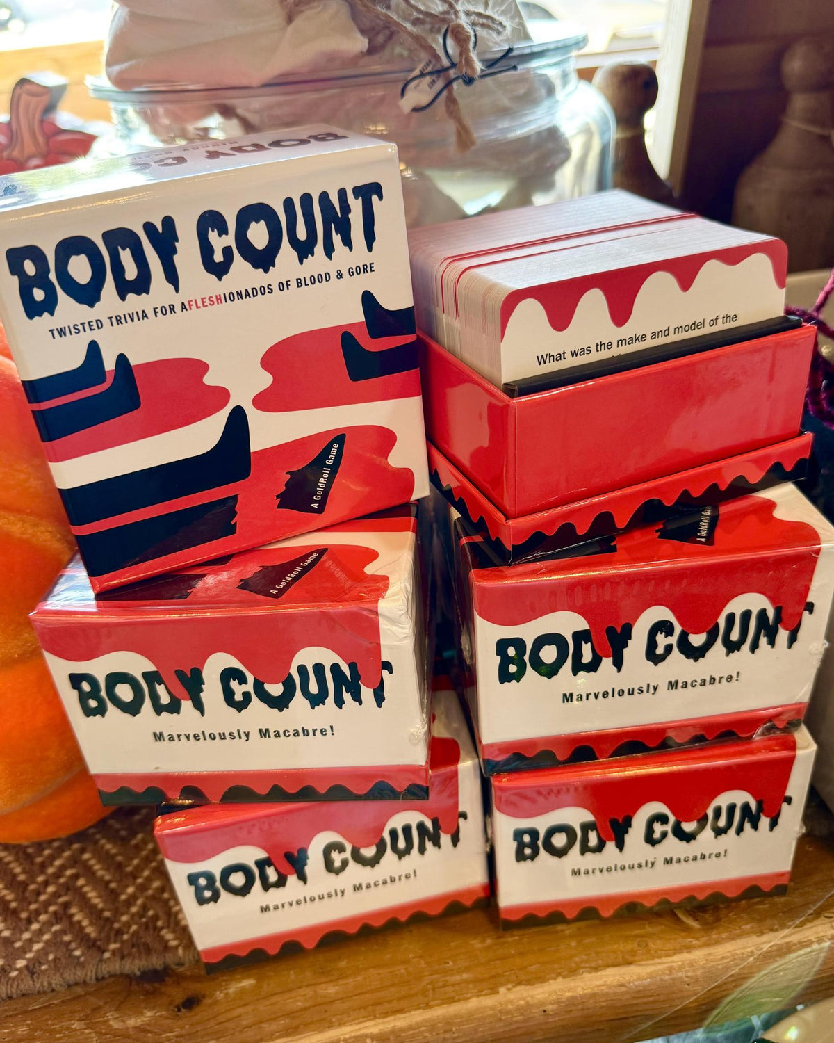A stack of 'Body Count' themed playing cards and matching boxes on a wooden surface. The packaging features a white background with bold red and navy blue graphics and text reading 'BODY COUNT' and 'Twisted Trivia for Allies of Blood & Gory'.