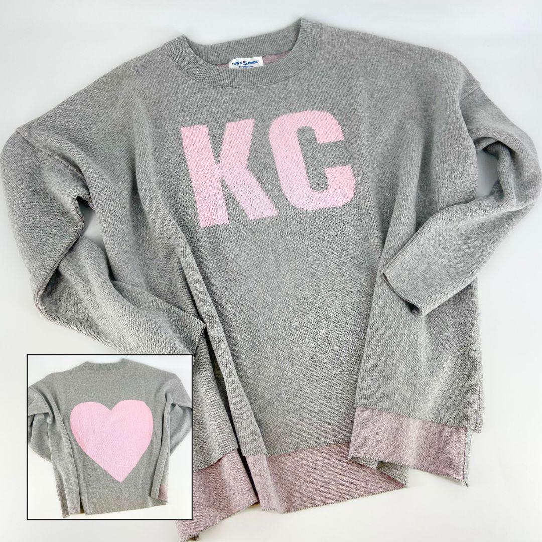 A pullover sweater with 'KC' text on the front and a heart symbol on the back, in grey and light pink colors, with a high-low hem and relaxed fit.