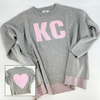 A pullover sweater with 'KC' text on the front and a heart symbol on the back, in grey and light pink colors, with a high-low hem and relaxed fit.
