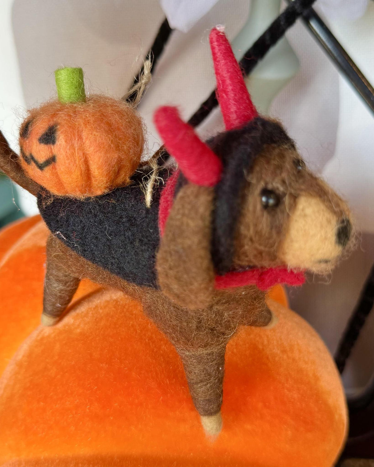 A small felted wool dog decoration wearing a black outfit and red horn hat, carrying an orange pumpkin on its back, set against an orange pumpkin base.