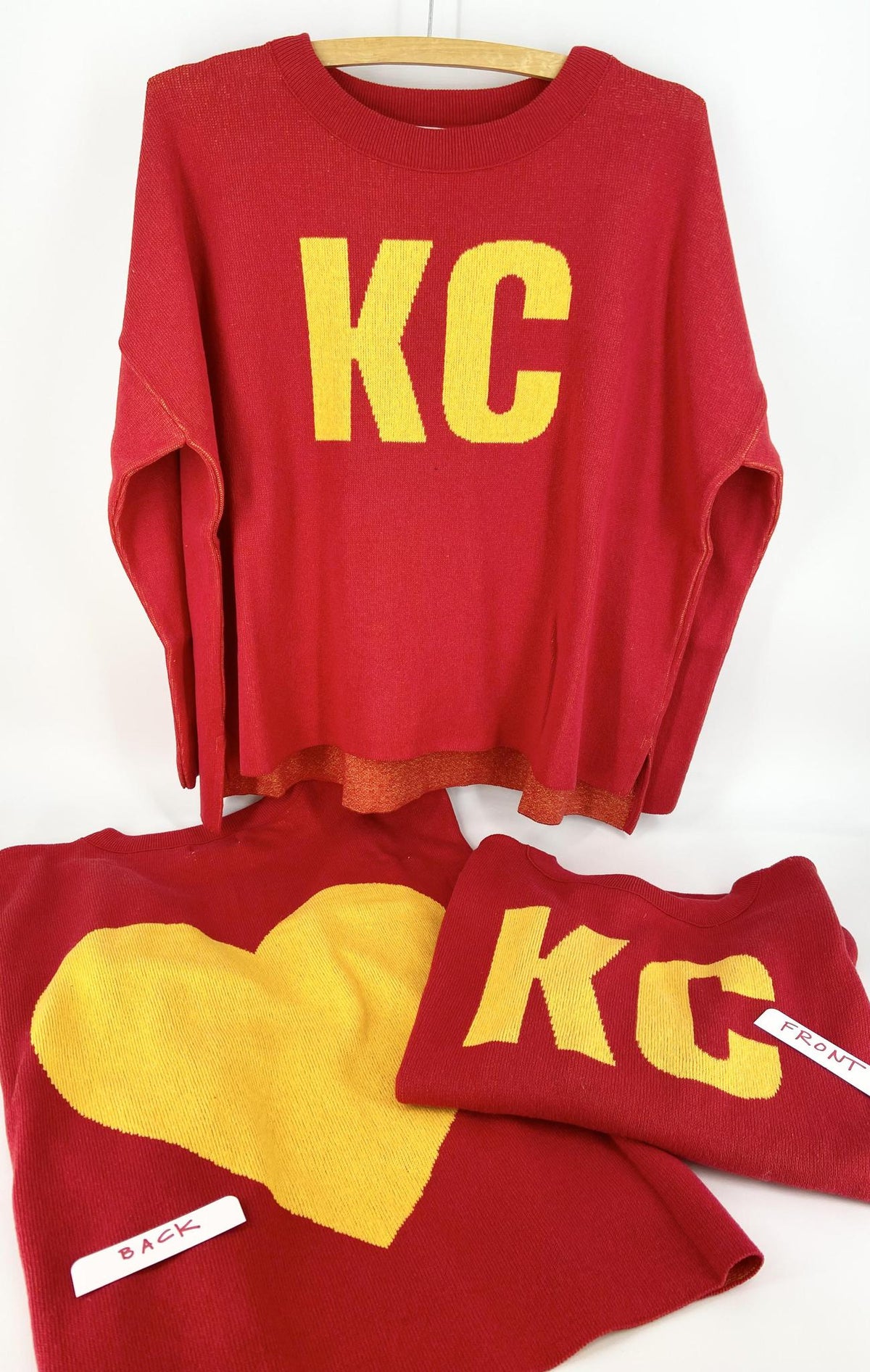 A red and gold pullover sweater with 'KC' text on the front and a heart shape on the back, displayed on a wooden hanger.