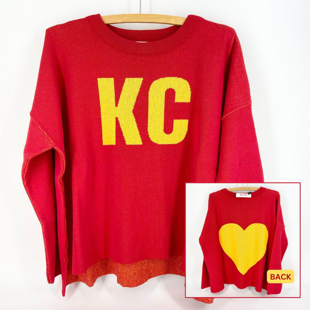 A red and gold pullover sweater with 'KC' text on the front and a heart shape on the back, displayed on a wooden hanger.