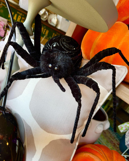 A large black spider decoration with long fuzzy legs, posed on a white and gray patterned pillow in a home setting with orange pumpkins.