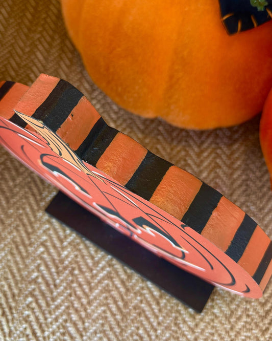 A close-up of a decorative Halloween sign shaped like a jack-o'-lantern, featuring orange and black stripes and a carved pumpkin face, displayed on a small black stand over a textured beige surface with pumpkins in the background.