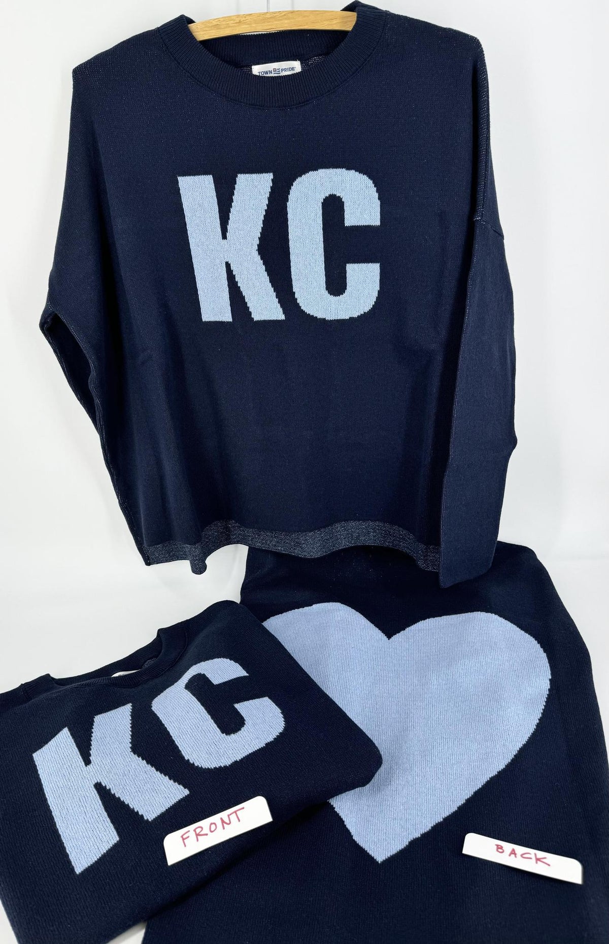 A navy pullover sweater with 'KC' written on the front and a heart shape on the back, displayed on a hanger.