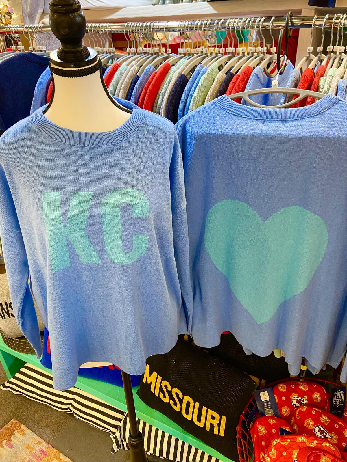 Two blue knit sweaters with 'KC' and heart designs on a mannequin in a store setting.