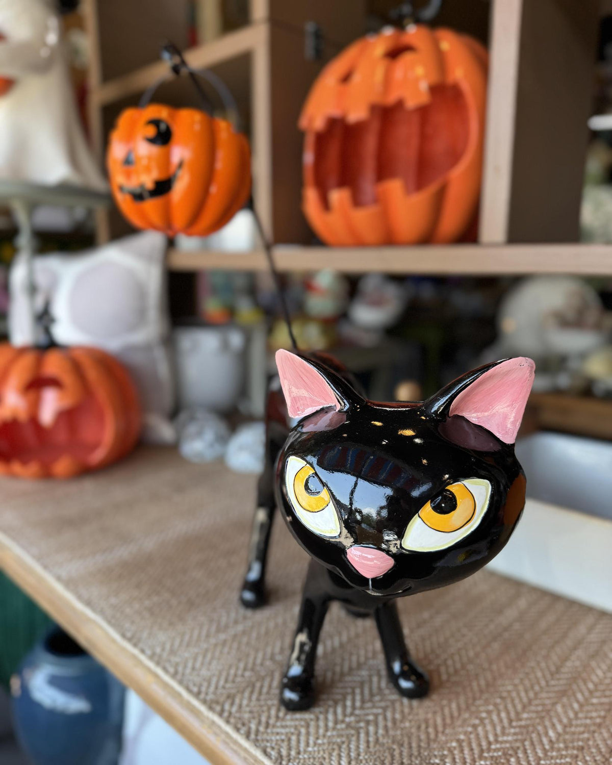 A glossy black cat figurine with pink ears and yellow eyes, styled on a shelf alongside orange jack-o'-lantern decorations.