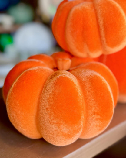 A close-up of a plush orange pumpkin decoration with a velvety texture, resting on a wooden surface with other similar pumpkins blurred in the background.